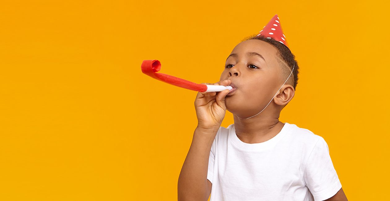 Happy boy celebrating a birthday, posing isolated at blank yellow wall, blowing whistle, Birthday hat on his head