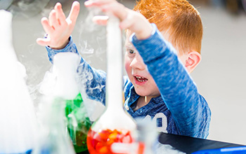 Boy playing with beakers with red and green liquid inside and smoke pouring out