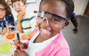 Girl smiling while holding a test tube with liquid inside. 2 other boys looking up at girl.  There are 2 beakers on the desk.