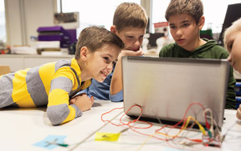 4 children looking at a computer with multicolored wires plugged in to a panel behind the computer.