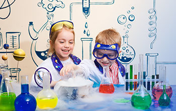 a boy and a girl wearing lab coats behind a desk with dry ice, beakers and tubes on it.