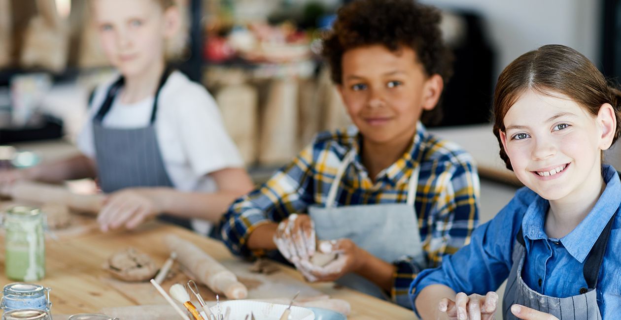 Happy little girl looking at you while sitting by table and making clay cup with her schoolmates on background