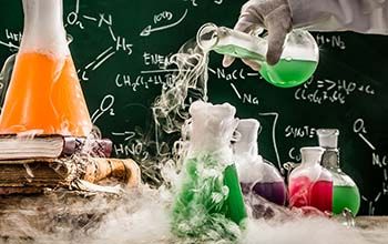 Hand pouring liquid into beaker on a desk.  Another beaker on a stack of books with smoke surrounding all beakers and table