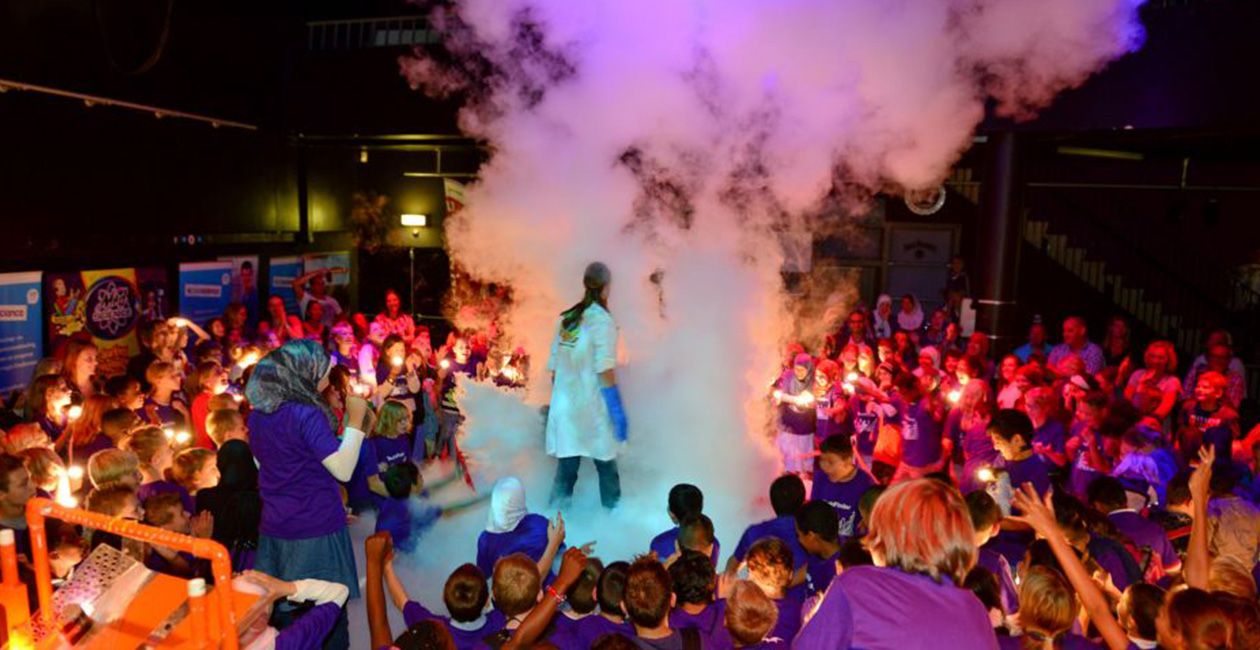 Mad scientist performing on stage surrounded by kids standing in a cloud of smoke