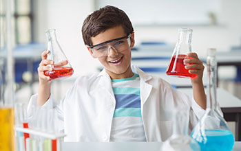 A boy in a white lab coat holding 2 flasks full of red liquid.